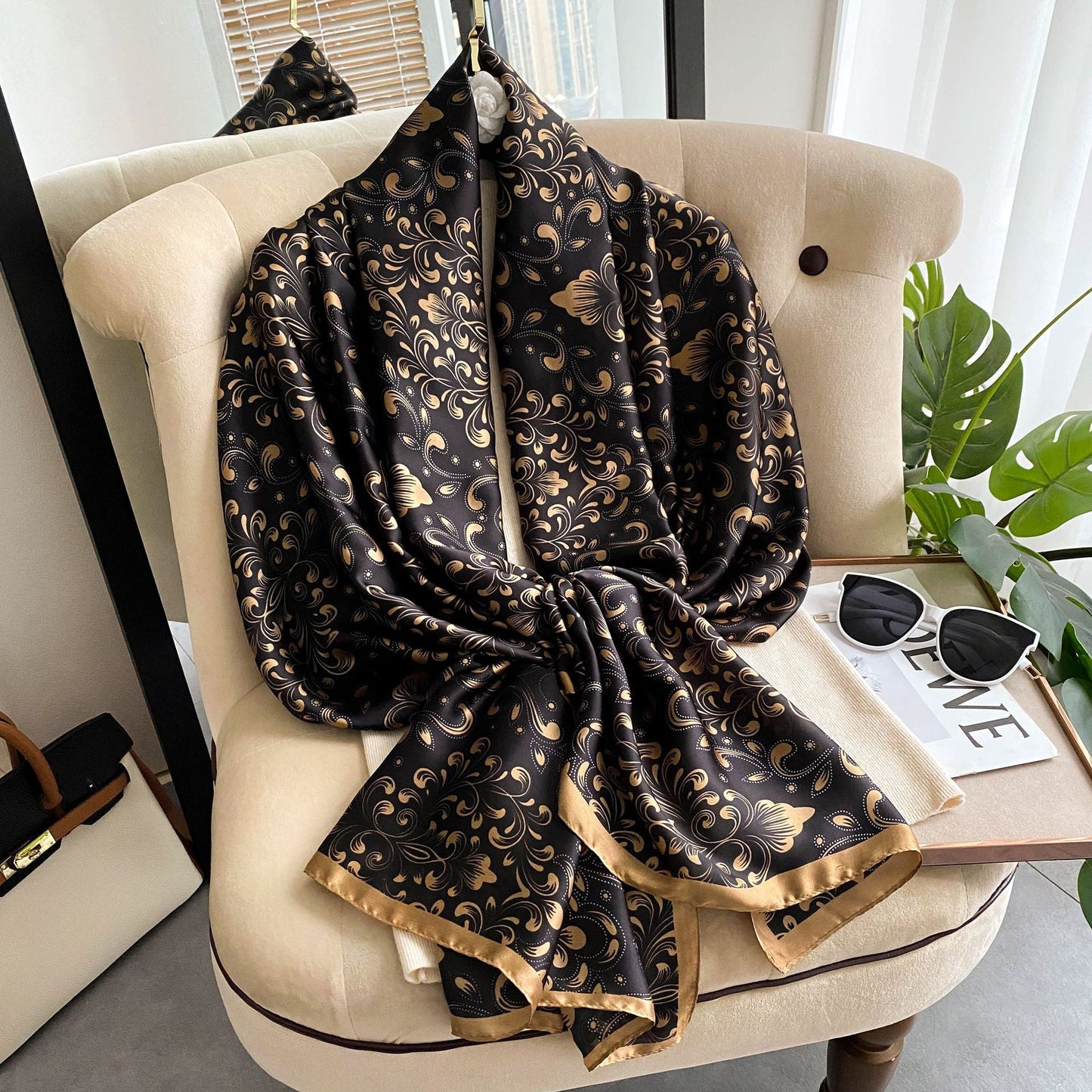 Black and gold patterned scarf draped over a beige chair with a plant and magazine in the background.